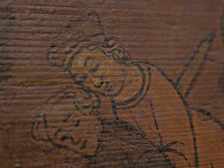 Dettaglio di una tavoletta lignea dipinta raffigurante una figura umana stilizzata, tracciata con linee scure su fondo marrone, con superficie segnata dal tempo.