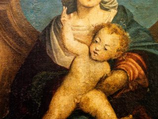 Dettaglio ravvicinato di un dipinto rinascimentale raffigurante la Madonna col Bambino. Si tratta della pala d’altare di Giovanni Antonio de’ Sacchis detto il Pordenone proveniente dalla Chiesa parrocchiale dei Santi Ilario e Taziano, nella frazione Torre. La Vergine, con capelli rossi ondulati e un'aureola dorata, indossa un manto blu scuro su una tunica chiara. È seduta e tiene in braccio il Bambino Gesù, che è nudo e in piedi sulle sue ginocchia, con il braccio sinistro sollevato e lo sguardo rivolto verso l'alto. Sullo sfondo si intravedono un cielo azzurro nuvoloso e, a sinistra, una struttura architettonica scura. La superficie del dipinto mostra segni di screpolatura del colore (craquelure).