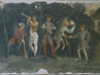 Un dettaglio di un affresco storico che raffigura un gruppo di uomini impegnati in una scena di festa e musica all'aperto, situata davanti a un fitto bosco scuro. Le figure indossano abiti rinascimentali, inclusi farsetti, calzamaglie colorate e cappelli tipici dell'epoca. A sinistra: Due musicisti suonano strumenti a fiato; uno è seduto e suona un lungo flauto dritto o una bombarda, l'altro è in piedi e suona una tromba sottile rivolta verso l'alto. Al centro: Un terzo musicista suona un flauto più piccolo, mentre accanto a lui un uomo è ritratto nell'atto di bere con vigore da una grande brocca bianca sollevata al viso. A destra: Un uomo in abiti azzurri osserva la scena con una mano sul fianco, vicino a quello che sembra l'ingresso di una struttura rurale o una piccola casa. Il dipinto presenta una tavolozza di colori terrosi e desaturati, con prevalenza di blu scuro, ocra, rosso mattone e grigio. La superficie è caratterizzata da evidenti segni di usura, crepe e mancanze di intonaco lungo i bordi, che testimoniano l'antichità dell'opera. Lo stile è narrativo e popolaresco, tipico dei cicli decorativi profani.