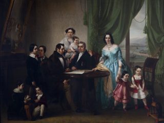 Un imponente ritratto di gruppo a olio dell'Ottocento ambientato in un interno borghese. La scena vede numerosi membri di una famiglia disposti attorno a un tavolo circolare in legno. Al centro: Un uomo siede al tavolo esaminando dei documenti, mentre un altro uomo in abito scuro siede di fronte a lui. Dietro di loro, una donna tiene in braccio un bambino piccolo. A destra: Una donna in un elegante abito azzurro lucido sta in piedi accanto a una finestra incorniciata da pesanti tende verdi. Tiene per mano una bambina piccola in abito rosa, mentre un'altra bambina in abito bianco e rosso sta in piedi vicino a una sedia. Dalla finestra si scorge un paesaggio costiero con un ponte e un treno a vapore in transito. A sinistra: Un gruppo di giovani uomini e ragazzi è disposto in piedi o seduto; in basso, due bambini giocano a terra con un piccolo cane. L'illuminazione è soffusa e proviene lateralmente, mettendo in risalto le diverse consistenze dei tessuti (seta, velluto e pizzo) e i dettagli degli arredi, come la carta da parati decorata e un grande specchio dorato sulla sinistra. L'atmosfera è formale e riflette la ricchezza e lo status sociale della famiglia rappresentata.