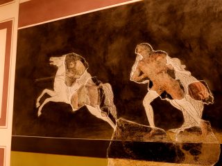 Dettaglio di una scena figurata su fondo nero che raffigura una amazzone su un cavallo bianco rampante e una figura maschile a piedi. Il pannello è una ricostruzione moderna che integra frammenti originali di intonaco antico, visibili per la loro texture materica e i colori più caldi rispetto alle sagome bianche di contorno.