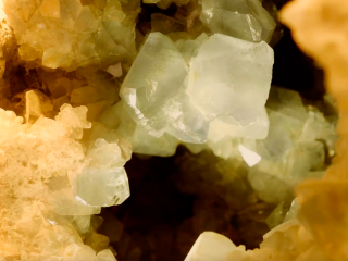 Un'inquadratura macro e ravvicinata dell'interno di un geode, focalizzata sulla crescita di grandi cristalli traslucidi. Al centro della composizione spiccano diversi cristalli romboedrici di grandi dimensioni, con superfici piatte e spigoli netti che riflettono la luce. Hanno un colore bianco ghiaccio. I cristalli più grandi sembrano emergere da una base irregolare di micro-cristalli di colore ocra e crema, che tappezzano le pareti della cavitàdel geode. L'illuminazione calda proviene dai lati, creando forti contrasti tra le facce brillanti dei cristalli e le zone d'ombra profonde (quasi nere) che si intravedono nel cuore del geode, dando un senso di profondità tridimensionale.