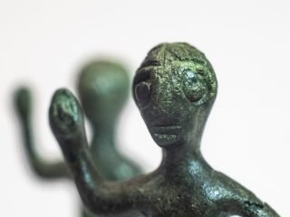 Primo piano ravvicinato del volto stilizzato e della testa di una statuetta in bronzo da Praturlone con patina verde-scuro. La messa a fuoco è sul viso, mettendo in evidenza le ampie orbite oculari e la texture rugosa della superficie metallica antica. Il volto è circondato da una capigliatura con riga centrale. Il braccio sollevato è parzialmente visibile e sfocato in primo piano, mentre lo sfondo è costituito da un'altra statuetta sfocata e da un fondo bianco neutro.