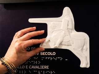 Mano che esplora un bassorilievo bianco raffigurante un cavaliere a cavallo, montato su un pannello con testo in caratteri braille.