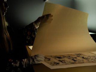 Il caveau si trova al piano interrato di Galleria Pizzinato. E' compostao da due stanze dedicate alla conservazione. Due stanze, una di consultazione e la seconda di conservazione. Entrambe con temperatura (16 gradi costanti) e umidità relativa controllate. Si tratta di sale in cui vengono conservate tavole, oggetti e riviste storiche dedicate al fumetto. L'immagine ritrae la curatrice museale al lavoro. In primo piano, due mani che indossano guanti bianchi di cotone sollevano con delicatezza un foglio di carta velina beige, translucida, posata sopra una tavola originale di fumetto inchiostrata. La tavola del fumetto, rivelata sotto la velina, è in bianco e nero e presenta quattro pannelli con scene illustrate e fumetti di testo (non leggibile). Nel primo pannello a sinistra, un gruppo di persone guarda verso l'alto; nel secondo, un uomo con indosso un abito cammina in una stanza d'ufficio; nel terzo, due personaggi parlano tra loro in piedi in una stanza; nel quarto, a destra, un uomo e una donna si guardano e sembrano conversare. Sullo sfondo a destra si intravedono le scatole di archivio scure con etichette. L'illuminazione è soffusa e focalizzata, proveniente dall'alto, lasciando il resto del soggetto e dell'ambiente in ombra. L'inquadratura è ravvicinata sulle mani e sui documenti.