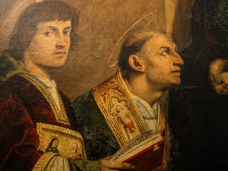 Dettaglio della pala d’altare di Giovanni Antonio de’ Sacchis detto il Pordenone. In primo piano sono raffigurati due santi: a sinistra, il più giovane ha capelli castani a caschetto guarda direttamente lo spettatore. Indossa una ricca veste damascata rosso scuro con bordi dorati decorati da piccole figure sacre. A destra, un santo più anziano con tonsura e aureola sottile è ritratto di profilo mentre guarda verso l'alto. Indossa paramenti liturgici verdi e oro e tiene in mano un libro aperto. La tecnica pittorica mostra un raffinato uso del chiaroscuro sui volti.