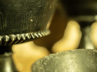 Primo piano estremo di due vasi in bucchero. La messa a fuoco è centrata sulla decorazione a rilievo del vaso superiore, che presenta una fila di piccoli dentelli semisferici lungo la carena e sottili incisioni verticali sopra di essi. In basso, il bordo frastagliato di un secondo vaso è visibile ma sfocato. Lo sfondo mostra forme calde e dorate fuori fuoco, illuminate da una luce soffusa.