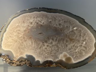 Un'inquadratura frontale di una sezione lucidata di un grande geode di giada. Il bordo esterno è costituito da una crosta rocciosa scura e grezza, seguita da sottili bande concentriche di colore grigio, bluastro e bianco che delimitano il perimetro. L'interno è riempito da una massa densa di cristalli di quarzo traslucidi, che degradano dal bianco opaco dei bordi verso un centro più chiaro. Al centro della sezione si trova una piccola cavità vuota, circondata da formazioni cristalline. La superficie è perfettamente levigata, tanto da riflettere la luce ambientale in modo uniforme. In basso a destra, sono visibili delle inclusioni circolari più scure che interrompono la regolarità delle bande. Il reperto poggia su un ripiano di vetro ed è illuminato da una luce diffusa contro uno sfondo grigio neutro, che mette in risalto la complessa geometria delle stratificazioni minerali. L'immagine evidenzia la perfezione delle formazioni geologiche stratificate nel corso di milioni di anni.