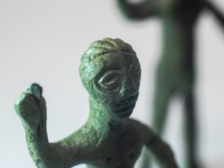 Macrofotografia del volto di un bronzetto da Praturlone. La messa a fuoco è centrata sugli occhi a mandorla, e sulla bocca, una sottile linea incisa sormontata dall'indicazione dei baffi. Il volto è incorniciato da una capigliatura all'indietro. La superficie metallica presenta una patina verde scuro molto materica, con evidenti porosità e segni del tempo. In primo piano, la mano destra sollevata della figura stringe il resto dell'asta di una lancia, ora perduta.