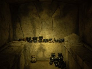 Fotografia dell'interno di una tomba a camera etrusca ricostruita. L'ambiente, dalle pareti irregolari e soffitto piatto, è illuminato frontalmente da una luce calda che rivela il corredo funebre. Su una banchina rialzata di fondo sono allineate numerose coppe e vasi in ceramica nera (bucchero), mentre sul pavimento in terra battuta sono raggruppati vasi di dimensioni maggiori e brocche. Ai lati si distinguono due letti funebri scolpiti nella pietra.