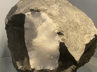 Inquadratura ravvicinata di un geode di natrolite: una roccia scura e opaca racchiude al suo interno una spettacolare cavità riempita di cristalli aciculari bianchi. Questi cristalli sottili, simili a aghi o a una peluria minerale, si irradiano verso l'esterno, creando una zona centrale chiara e traslucida dalla texture setosa. La roccia esterna è di colore grigio scuro, con una superficie irregolare, spigolosa e frammentata, che contrasta nettamente con la delicatezza dei cristalli interni. La luce colpisce direttamente la sezione cristallina, facendola brillare e mettendo in risalto la fitta trama degli aghi minerali.