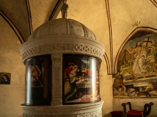 Veduta ravvicinata di un antico fonte battesimale monumentale in pietra bianca, situato all'interno di una chiesa o cappella con soffitto a volta. La struttura è cilindrica e sormontata da una cupola in pietra decorata con motivi vegetali a rilievo e una piccola statua sulla sommità. Il corpo centrale del fonte ospita una serie di tavole dipinte centinate che seguono la curvatura della struttura. Sono chiaramente visibili due dei pannelli analizzati in precedenza: a sinistra il Battesimo di Cristo e al centro il Banchetto di Erode (con Salomè che regge il vassoio con la testa del Battista). Le tavole fungono da ante di chiusura per il vano interno del fonte. Sulla parete di fondo a destra, all'interno di un'arcata cieca, è presente un grande affresco della Resurrezione di Cristo, che mostra Gesù che emerge dal sepolcro con il vessillo crociato, circondato dai soldati romani dormienti. L'ambiente è sobrio, con pareti intonacate e arredi liturgici minimi, tra cui una sedia a forchetta in legno scuro con cuscino rosso.