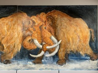 Un dipinto murale a tre pannelli, in stile espressionista, che raffigura due grandi mammut lanosi con folta pelliccia color ocra-marrone che si scontrano con le zanne, posizionati su un terreno ghiacciato e con uno sfondo scuro. I mammut sono raffigurati con pennellate audaci e strutturate. Il mammut a sinistra è leggermente più avanti, mentre il mammut a destra si sta appoggiando. Le loro lunghe zanne bianche sono incrociate al centro, e spruzzi di neve e ghiaccio si alzano dove si incontrano. L'opera è firmata "Alberto Magri" nell'angolo in basso a destra. Il dipinto è esposto su un muro bianco con modanature decorative nella parte inferiore.
