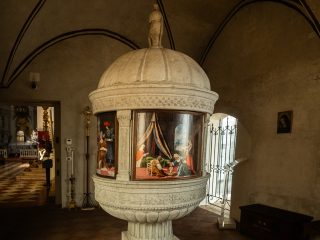 Fonte battesimale in pietra con cupola scolpita, decorata da pannelli dipinti, collocata all’interno di una chiesa storica con volte ad arco e luce naturale laterale.