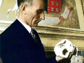 Un ritratto storico del Conte Giuseppe di Ragogna, raffigurato di profilo mentre indossa un abito scuro formale e una cravatta. Il Conte tiene tra le mani e osserva con attenzione due frammenti di pietra. Sullo sfondo, una parete ospita un grande stemma araldico decorato con motivi a rombi rossi e grigi. L'immagine ha un'atmosfera solenne e documentaristica, tipica degli studi archeologici del passato.