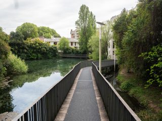 "Una passerella pedonale in legno e metallo attraversa un corso d'acqua immerso nel verde in una zona residenziale. Il sentiero, fiancheggiato da balaustre scure, si sviluppa in linea retta sopra lo specchio d'acqua, circondato da alberi rigogliosi e vegetazione spontanea. In lontananza, oltre gli alberi, si intravedono alcuni edifici residenziali, creando un contrasto tra l'ambiente urbano e l'oasi naturale."