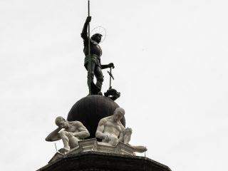 Scultura monumentale vista dal basso: una figura in piedi sopra una sfera tiene un’asta, mentre due figure sedute sono poste alla base del monumento.