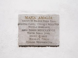 "Una targa commemorativa in pietra chiara affissa su una parete bianca intonacata. La targa presenta un'iscrizione in latino incisa, che inizia con le parole 'MARIA AMALIA' in caratteri prominenti, seguita da diverse righe di testo commemorativo in maiuscolo."