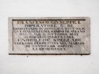 "Una lapide commemorativa in marmo chiaro affissa su una parete bianca. L'iscrizione, incisa in caratteri maiuscoli, ricorda che l'imperatore Francesco Giuseppe I soggiornò in questa casa dal 7 all'11 ottobre 1852, in occasione di simulazioni di battaglie della cavalleria nel campo di Aviano. La targa è stata posta dalla famiglia nobile De Spelladi per tramandarne il ricordo."