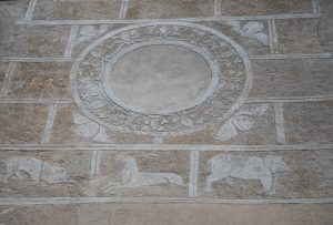 Dettaglio di un graffito esterno su Palazzo Marone a Pordenone, raffigurante una decorazione geometrica a riquadri: al centro un grande tondo inciso con motivi vegetali e circondato da testine di cherubini angolari, mentre nel registro inferiore sono visibili sagome di animali, tra cui un cervo e un suide, incise nella malta.