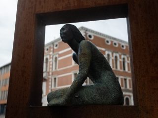 "Una scultura in bronzo raffigurante una figura femminile seduta, con il corpo caratterizzato da una texture rigata e il volto rivolto verso l'alto. La statua è inquadrata attraverso una cornice metallica squadrata color ruggine. Sullo sfondo, leggermente fuori fuoco, si scorgono gli edifici storici che si affacciano su Piazza Cavour a Pordenone."