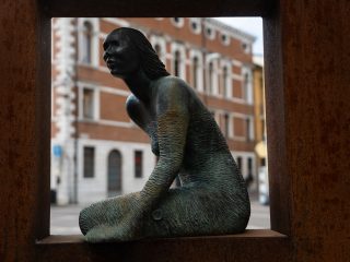 "Un'inquadratura ravvicinata di una scultura in bronzo che ritrae una figura femminile in posa seduta, con la testa rivolta verso l'alto e la superficie del corpo lavorata con una texture finemente rigata. La statua è collocata all'interno di una cornice metallica squadrata color ruggine che funge da quinta scenica. Sullo sfondo, sfuocati, si distinguono l'architettura classica in mattoni e le finestre di un palazzo storico che si affaccia su Piazza Cavour a Pordenone."