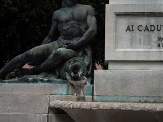 "Dettaglio di un monumento commemorativo in pietra e bronzo. Sulla sinistra, una statua bronzea di una figura maschile nuda, in posa seduta, poggia su un basamento di pietra; la figura sembra versare acqua da un recipiente a forma di anfora verso una vaschetta sottostante. Sulla destra, una parte del basamento in pietra riporta l'iscrizione 'AI CADUTI' seguita dai numeri romani 'MCMX' e 'MCMX' (riferimento alla Prima Guerra Mondiale). La pietra mostra segni di ossidazione verdastra tipica dell'esposizione agli elementi."
