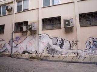 Murale in bianco e nero su un edificio: una grande figura distesa si estende lungo la parete, con graffiti e finestre con grate sopra l’opera.