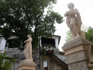 "Una veduta in un cortile o giardino storico caratterizzato da elementi architettonici e scultorei in pietra. In primo piano, due statue in pietra su alti basamenti: a sinistra una figura femminile in piedi e a destra una figura maschile (probabilmente allegorica, che tiene un oggetto sferico). Sullo sfondo, un edificio antico con una scalinata esterna in pietra che conduce a una loggia coperta, circondato da vegetazione e un grande albero. L'atmosfera è quella di un luogo ricco di storia e fascino architettonico."