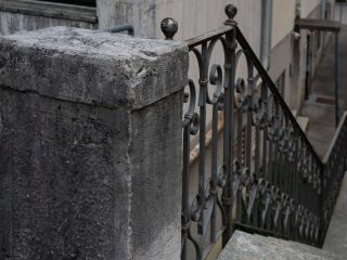 "Un dettaglio architettonico che mostra un solido pilastro in pietra grigia, dall'aspetto ruvido e segnato dal tempo, che fa da sostegno a una ringhiera in ferro battuto. La ringhiera è caratterizzata da motivi a volute e termina con un piccolo pomello decorativo sferico. Sullo sfondo, si intravede una scalinata che scende verso un passaggio esterno, suggerendo un contesto urbano o di un edificio storico."