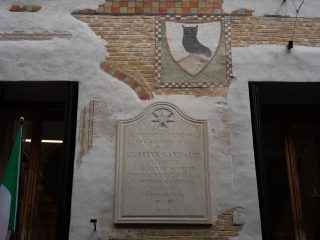 "Una lapide commemorativa in pietra chiara affissa sulla facciata di un edificio antico, caratterizzata da intonaco parzialmente scrostato che rivela mattoni a vista e affreschi decorativi, tra cui uno stemma raffigurante un gufo. L'iscrizione sulla targa recita: 'A RICORDO PERENNE DEL 2 MARZO 1867 IN CUI GIUSEPPE GARIBALDI OSPITE ALLE LOTTE SUPREME CONTRO I NEMICI D'ITALIA IL POPOLO COMMOSSO INCITAVA I PORDENONESI P.P. 1882'. La parte superiore della targa è decorata con una stella scolpita. A sinistra della lapide è visibile parte di una bandiera italiana."