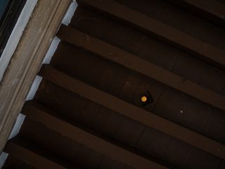 Soffitto in legno con travi parallele scure, visto in diagonale dal basso; un piccolo elemento circolare è visibile tra le travi.