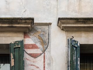 "Dettaglio di una facciata di un edificio antico con intonaco chiaro e due finestre affiancate, dotate di persiane in legno verde visibilmente scrostate. Tra le due finestre è dipinto uno stemma araldico raffigurante un leone rampante dorato su sfondo grigio nella metà superiore, e bande orizzontali rosse e bianche nella metà inferiore."