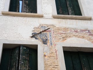 "Dettaglio della facciata di un edificio antico con quattro finestre dotate di persiane in legno verde scrostate. Tra le due finestre al piano inferiore è visibile un frammento di affresco o decorazione pittorica parzialmente erosa, che mostra motivi geometrici e una figura scura, attraversata in diagonale da un sottile tirante metallico."