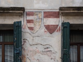 "Un dettaglio della facciata di un edificio storico, caratterizzato da intonaco in parte scrostato. Al centro, tra due finestre con persiane in legno verde scuro, sono dipinti due stemmi araldici: quello di sinistra presenta bande orizzontali alternate e tre gigli, mentre quello di destra mostra la classica fascia orizzontale rossa e bianca. L'insieme riflette l'aspetto tipico dell'architettura d'epoca in contesti urbani, con i segni dell'usura del tempo che ne arricchiscono il valore storico."