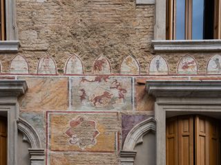 "Dettaglio della facciata di un edificio storico in mattoni a vista, caratterizzato da decorazioni pittoriche su vari livelli. Nella parte superiore, un fregio composto da una serie di piccole arcate ospita figure allegoriche e simboli, tra cui creature mitologiche e segni zodiacali. Al centro si trova uno stemma araldico raffigurante un leone rampante, mentre nella parte inferiore sono visibili riquadri geometrici dipinti con motivi floreali. Ai lati della composizione si aprono finestre con cornici in pietra e persiane in legno."