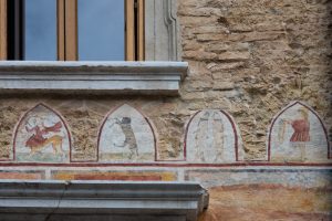 Dettaglio di un fregio a fresco dipinto sotto un cornicione in pietra del Palazzo Marone a Pordenone. Quattro formelle ogivali separate da cornici rosse si susseguono su una parete in muratura grezza: la prima raffigura una Centauressa con arco e frecce, la seconda un orso che si arrampica, la terza due figure stilizzate di pesci affrontati e la quarta una figura umana con un berretto.
