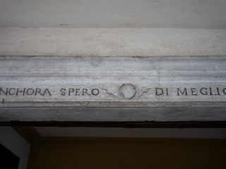 "Un dettaglio architettonico che mostra un architrave in pietra grigia, decorato con un'iscrizione incisa in caratteri capitali neri che recita 'ANCHORA SPERO DI MEGLIO'. Al centro, tra le due parti del testo, è presente un piccolo disco circolare inciso nella pietra. L'architrave sovrasta un'apertura, probabilmente l'ingresso di un edificio storico."