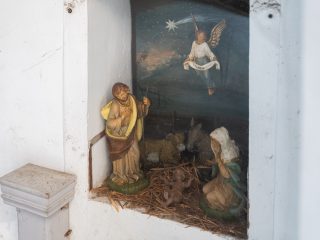 Presepe allestito in una nicchia: statue attorno a una mangiatoia con animali, una figura alata sospesa sopra la scena e una stella sullo sfondo dipinto.