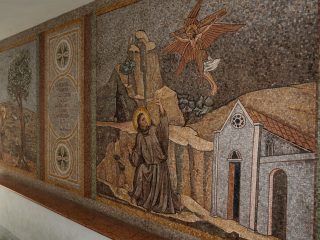 Mosaico murale con una scena religiosa: figure in abiti monastici in un paesaggio collinare, una figura alata sopra di loro e un edificio sacro sul lato destro.