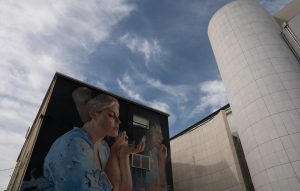 "Un grande murales dipinto sulla parete scura di un edificio, che raffigura una donna intenta a truccarsi mentre si guarda in un piccolo specchio. Indossa un indumento azzurro decorato con motivi di volatili. Accanto alla figura principale si nota l'immagine pi&ugrave; tenue di una persona che sembra compiere lo stesso gesto. Alla destra dell'edificio si staglia una struttura moderna caratterizzata da un'alta torre cilindrica rivestita di piastrelle bianche quadrate, il tutto sotto un cielo azzurro con nuvole sparse."