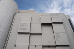 "Una facciata architettonica moderna e minimalista di un edificio di colore chiaro, caratterizzata da una serie di volumi geometrici sporgenti a forma di parallelepipedo. Su tre di questi pannelli sono incise delle poesie in lingua italiana. Sul lato sinistro &egrave; visibile parte di una struttura cilindrica moderna rivestita di piastrelle bianche, il tutto sotto un cielo azzurro con alcune nuvole."