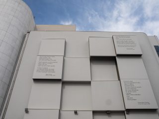‘A modern, minimalist façade of a light-coloured building, characterised by a series of protruding geometric volumes in the shape of parallelepipeds. Poems in Italian are engraved on three of these panels. On the left-hand side, part of a modern cylindrical structure clad in white tiles is visible, all set against a blue sky with a few clouds.’