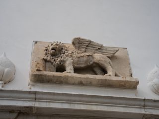"Scultura in rilievo in pietra raffigurante il Leone di San Marco, alato e rivolto verso sinistra, montata su una parete bianca. La figura del leone è fiancheggiata simmetricamente da due elementi decorativi in pietra dalla forma arrotondata, che ricordano dei boccioli o delle pigne."
