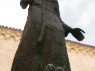 "Ripresa dal basso e fuori fuoco di una statua in bronzo raffigurante un frate in abito francescano con le braccia tese, in un gesto di accoglienza. In primo piano si intravedono, anch'essi sfocati, dettagli materici della scultura, mentre sullo sfondo è visibile la facciata in mattoni di un edificio storico caratterizzata da archetti decorativi."