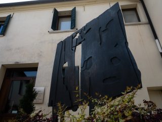 Un'opera scultorea contemporanea in metallo scuro dalle forme geometriche traforate, posizionata in un cortile davanti a un edificio dall'architettura sobria con persiane verdi. La scultura ha un aspetto imponente e organico, con aperture irregolari che ricordano sagome umane o forme naturali.
