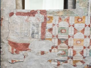 "Un dettaglio ravvicinato di una facciata storica decorata con affreschi antichi, visibilmente usurati dal tempo. Sulla sinistra, una sezione mostra tracce di un disegno architettonico, mentre sulla destra la superficie è ricoperta da un motivo geometrico regolare e ripetitivo, composto da quadrati e triangoli nei toni del rosso, ocra e verde, arricchiti da cerchi centrali. L'intonaco presenta crepe e zone scrostate, che lasciano intravedere la texture sottostante, a testimonianza dell'antichità e del pregio artistico dell'edificio."