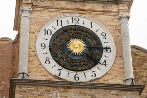 Dettaglio di un antico orologio astronomico posizionato su una facciata in mattoni a vista, affiancato da due colonne in pietra. Il quadrante principale presenta una numerazione classica da 1 a 12 in cifre romane. Al centro, un disco rotante pi&ugrave; piccolo mostra il sole dorato raggiante circondato da un anello con i segni dello zodiaco e i nomi dei mesi in italiano arcaico, incisi lungo la circonferenza. Le lancette scure indicano l'ora, mentre il meccanismo centrale illustra il moto apparente dei corpi celesti.