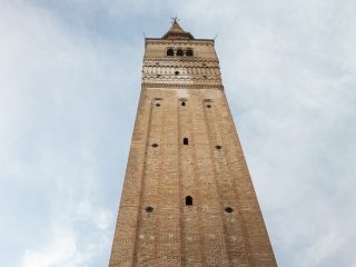 "Una veduta dal basso verso l'alto di un antico e slanciato campanile in mattoni a vista. La struttura si sviluppa verticalmente con un design austero, caratterizzato da lesene che corrono lungo tutta l'altezza, ritmate da piccole aperture ad arco e piastre circolari in metallo che fungono da tiranti strutturali. Nella parte superiore, il campanile presenta una cella campanaria con trifore e termina con una cuspide piramidale sormontata da una croce. Il campanile si staglia contro un cielo azzurro leggermente velato, con l'angolo di un edificio color ocra visibile nell'angolo in basso a sinistra e un frammento di tetto sporgente nell'angolo in alto a destra."