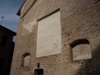 "Una veduta in prospettiva di una parete esterna di un edificio antico costruito in mattoni a vista. La facciata è caratterizzata da una grande porzione centrale intonacata di bianco, di forma rettangolare, circondata dal mattone irregolare. Ai lati e in basso sono presenti nicchie murate con archi a tutto sesto, anch'esse intonacate. Sulla sinistra si intravede l'angolo di un altro edificio adiacente sotto un cielo terso. L'aspetto complessivo è quello di una struttura storica sottoposta a restauri o modifiche nel corso del tempo."